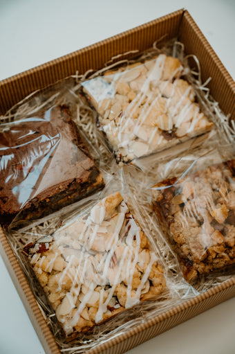 Box of assorted baked goods including brownies and crumbly bars with white drizzle on a light background.